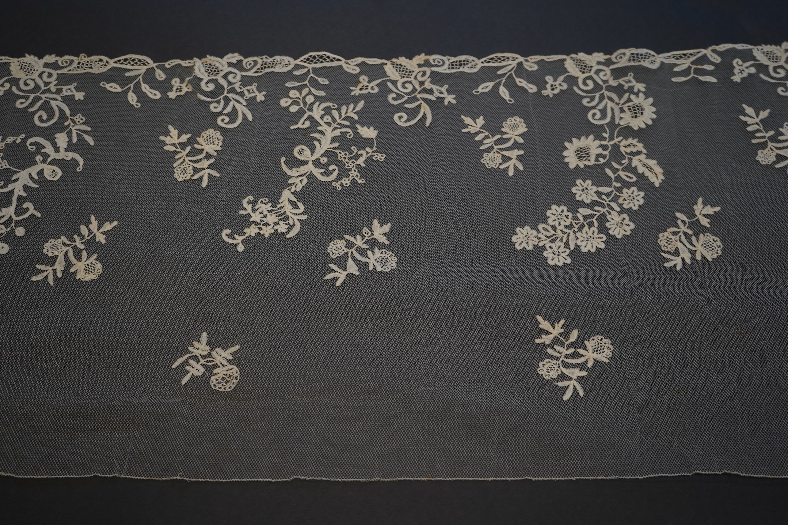 A 19th century five yard flounce of Brussels bobbin appliqué lace applied onto fine machine net, worked in a trailing floral design with an ornate border and sprig motifs, 457.5 cm long x 36cm deep. Condition - possibly